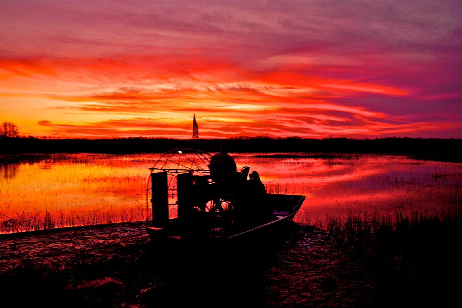 An airboat silhouetted against a red and purple sunset over a marsh, the kind of boat used on the grassy shallows of Glades County, Florida