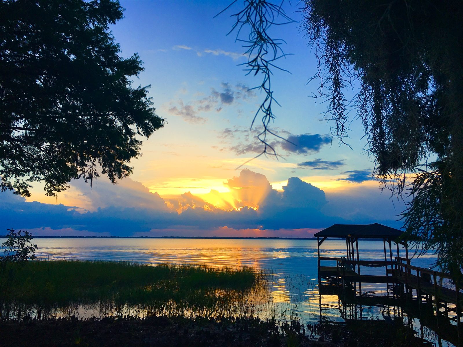 Sunset over a wide freshwater lake with reeds and a covered boat dock framed by moss-draped trees